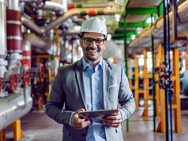 Man in a hard hat and business suit holding a tablet and smiling in front of factory pipes.