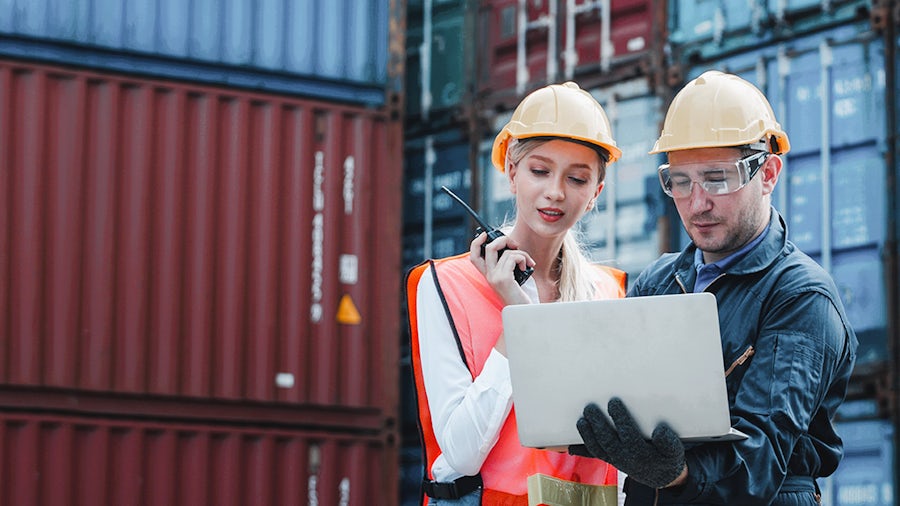 Two workers in safety gear stand in front of shipping containers, one holding a radio and the other using a laptop.