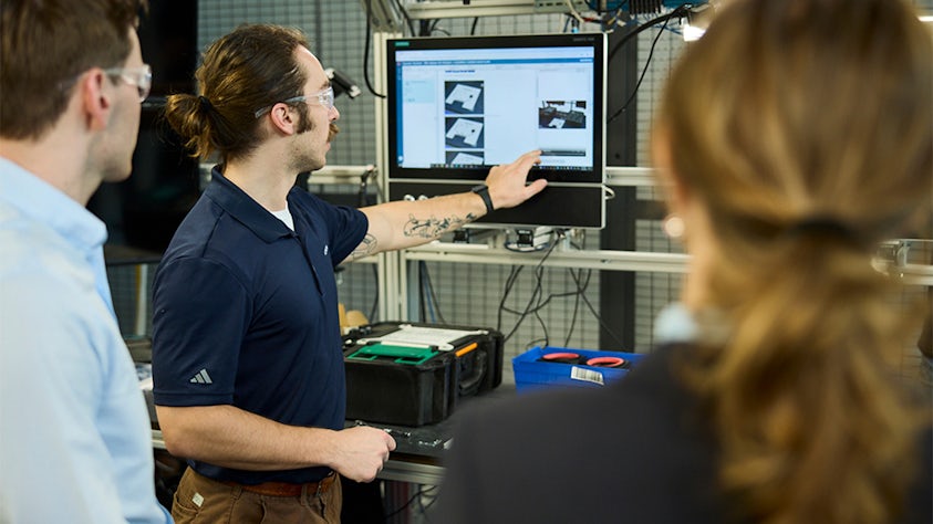 Three individuals in a technical or lab setting, gathered around a computer screen displaying images and text, with various equipment and tools on the table.