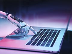 A robotic hand presses a key on a laptop computer keyboard.