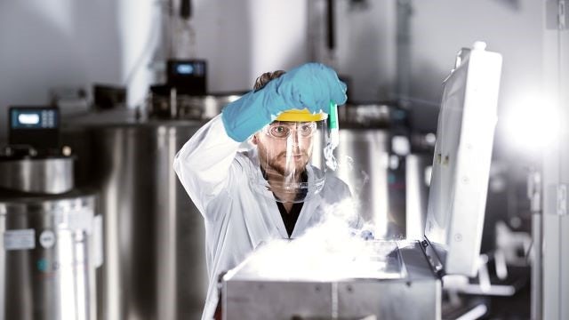 A man wearing a blue glove and face mask handles a deep-frozen glass vial. 