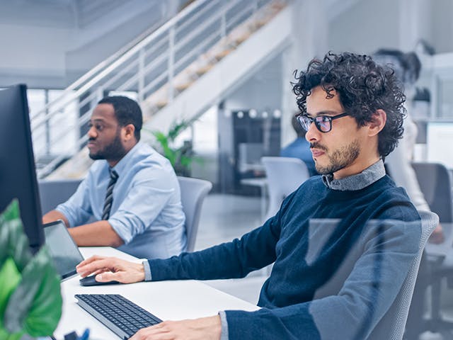 Two professionals working at their desks in a modern office environment. The man in the foreground, wearing glasses and a dark sweater, is focused on his computer screen, while the other man in business attire is engaged with his laptop. The office has an open layout with bright lighting, contemporary furniture, and a staircase in the background.