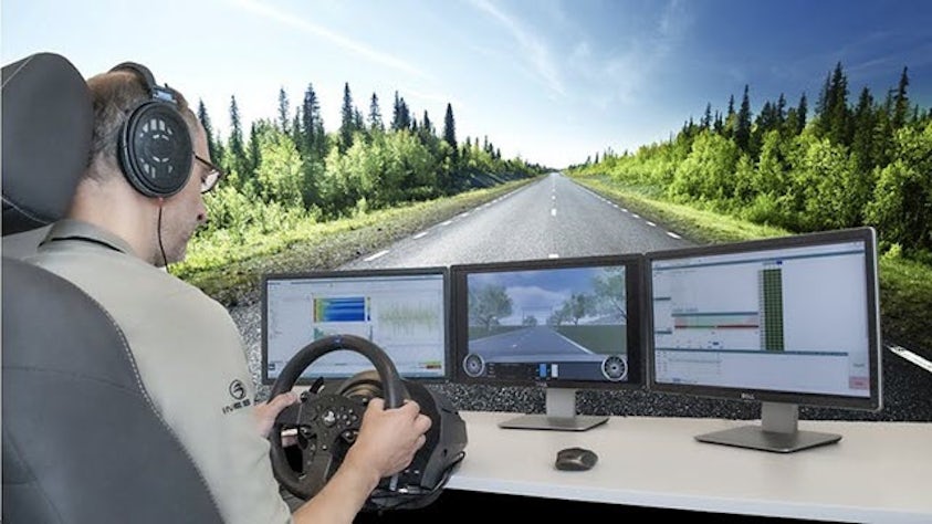 Engineer using driving simulator with triple-monitor setup and steering wheel controls, testing vehicle dynamics against scenic road background.