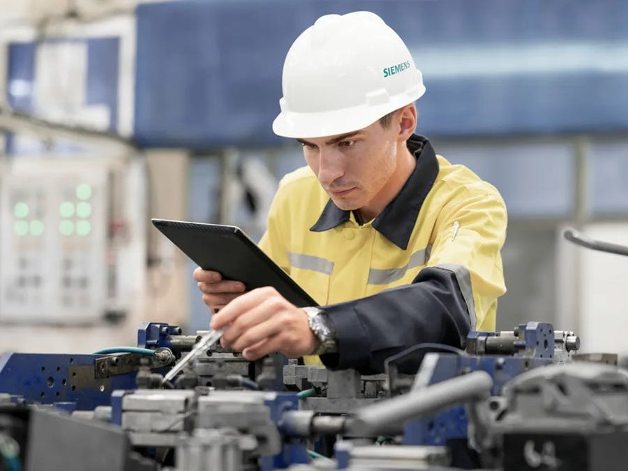 Tecnico Siemens con elmetto di sicurezza bianco e giubbotto giallo controlla macchinari industriali utilizzando un tablet in ambiente di produzione.
