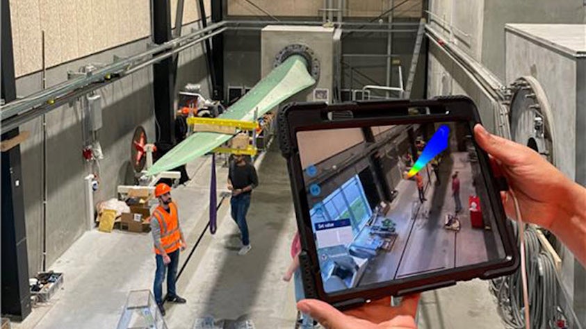 Engineer using tablet with AR technology to monitor wind tunnel testing, displaying digital overlay of aerodynamic data on physical aircraft model setup.