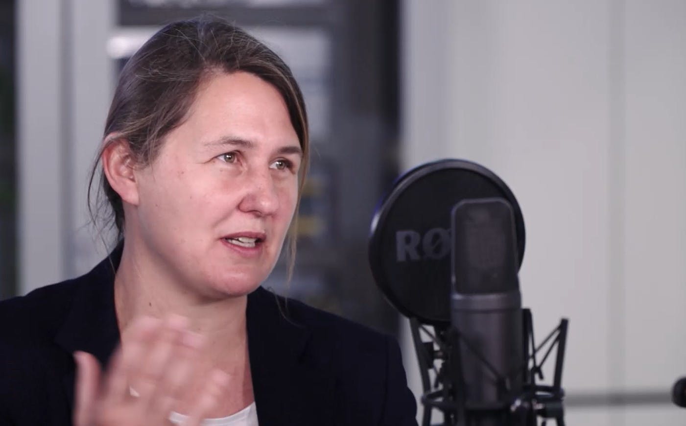 A person speaking into a professional microphone setup in what appears to be a podcast or recording studio environment, wearing a black blazer and light-colored top.