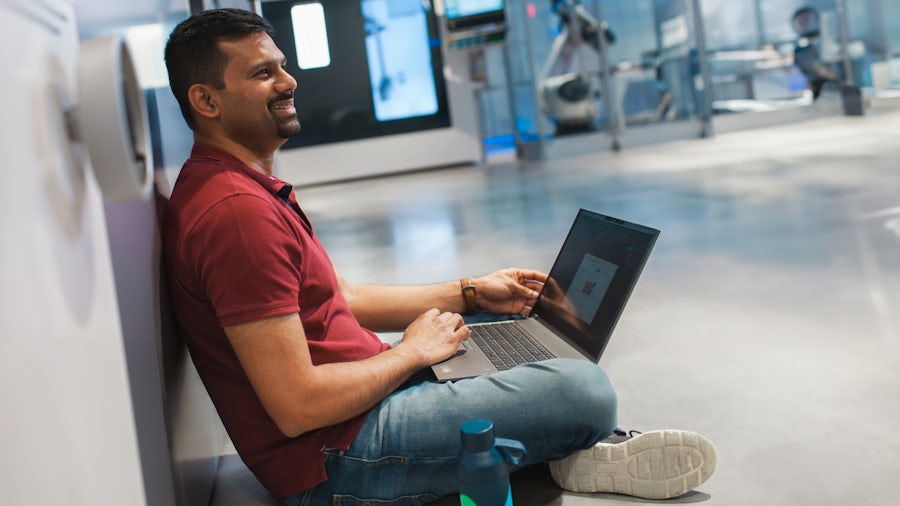 An engineer working on his laptop while sitting on the floor of a manufacturing facility.