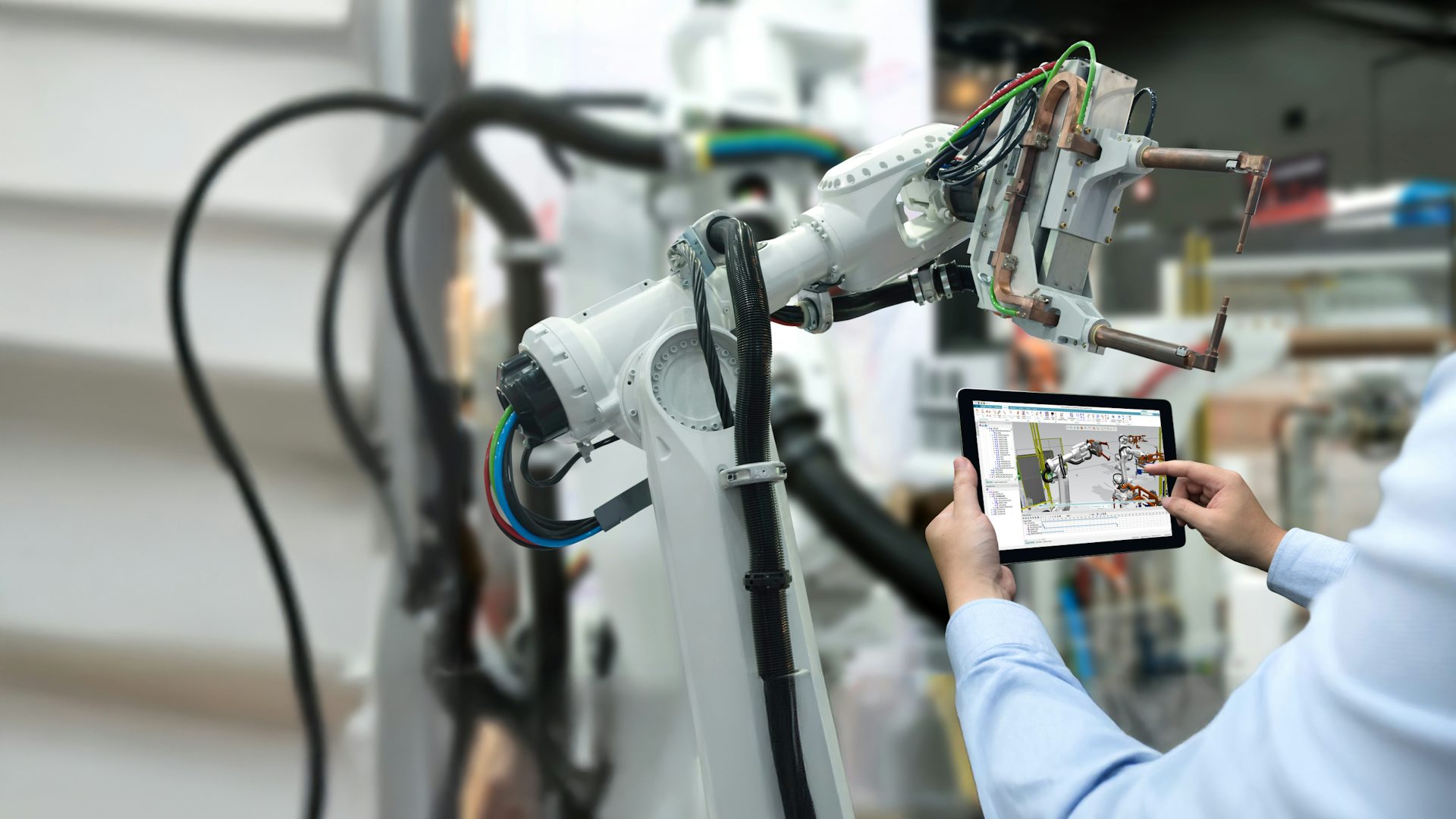 Manufacturing engineer reviewing a robotics digital twin model in Tecnomatix Process Simulate software on a tablet in front of the real robotic system.