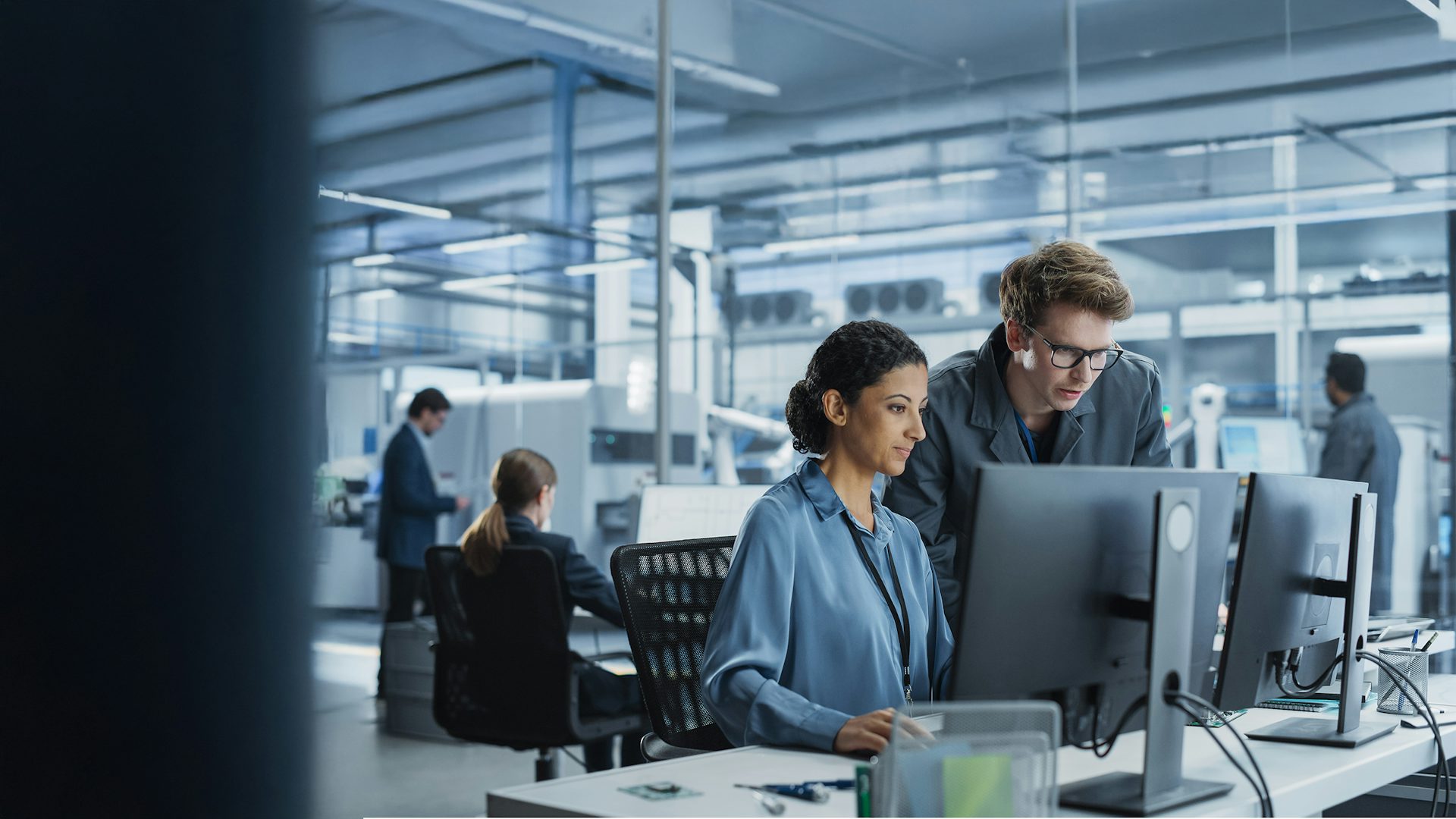 Engineers collaborating on a project using a computer in a factory with robotic arms producing devices for the tech industry.
