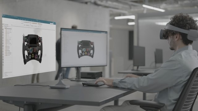 Ingeniero que utiliza un ordenador de sobremesa con un monitor virtual a la izquierda para ingeniería inmersiva con el casco de realidad virtual Sony XR y NX Immersive Designer. Ambos monitores muestran una ventana de NX con un diseño CAD del volante de un coche de F1.
