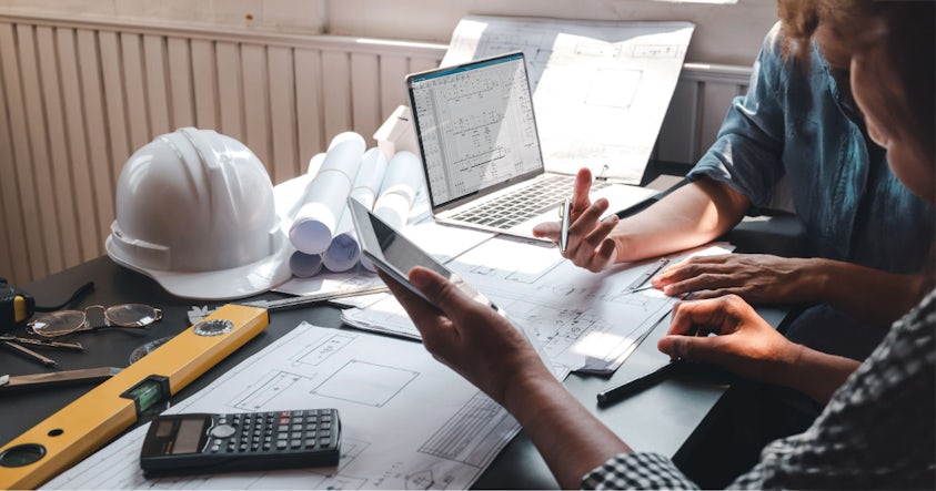 Two engineers engaged in a project discussion at a table, equipped with a laptop and electrical schematics drawing.
