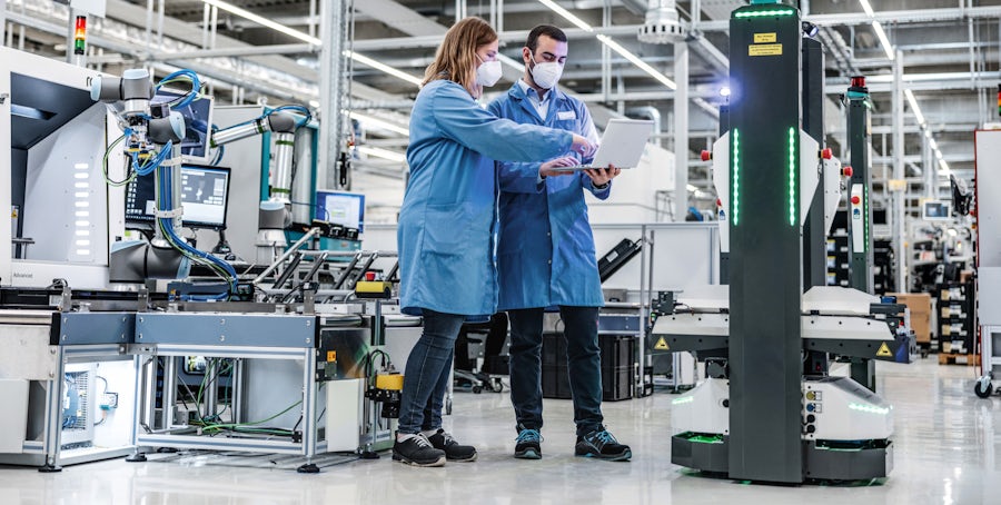 Two manufacturing operations managers reviewing materials management and intralogistics plans on a laptop in a manufacturing facility near AGVs