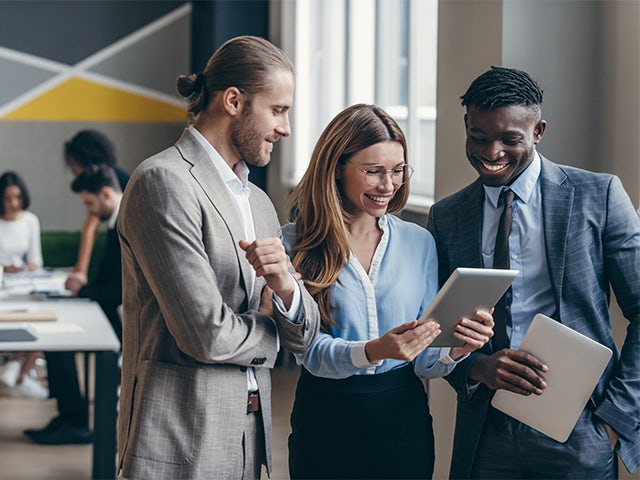 Three young business people looking at digital tablet while their colleagues are working behind them.