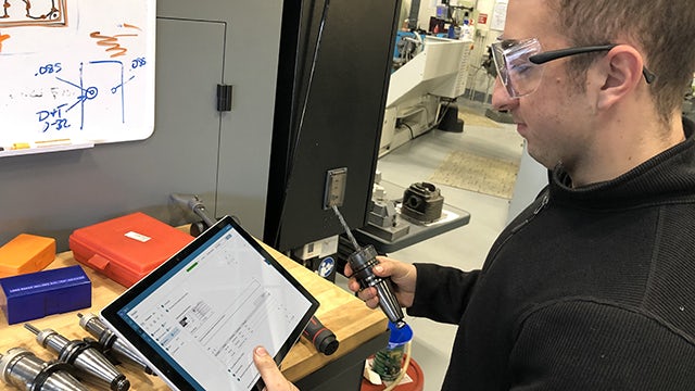 Male in a machine shop setting. He is wearing safety goggles and a black fleece. He's holding the tool in one hand and a tablet in the other. The tablet has instructions on the screen.