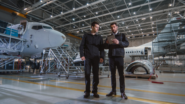 Two male maintenance mechanics look at a tablet while they're standing in an airplane hangar. 