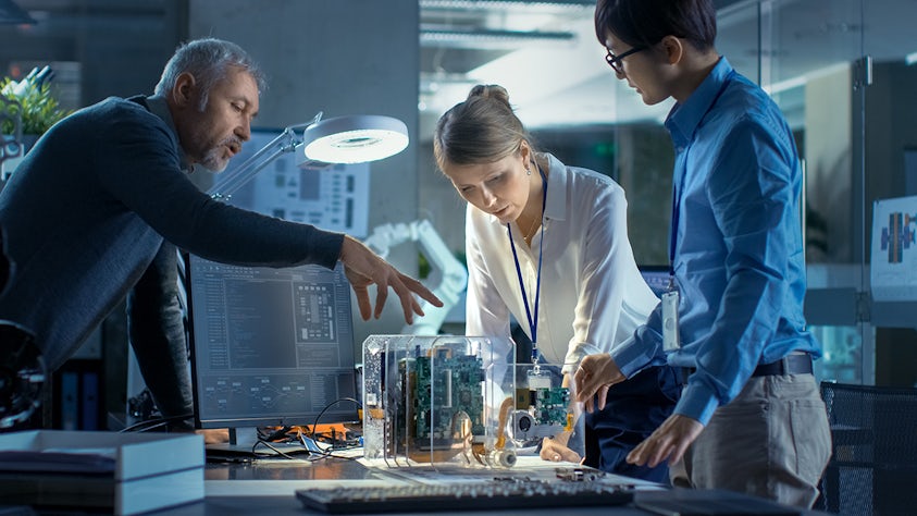 Team of computer engineers at a desk selecting printed circuit boards, with programming displayed on a computer screen in a high-tech lab setting.