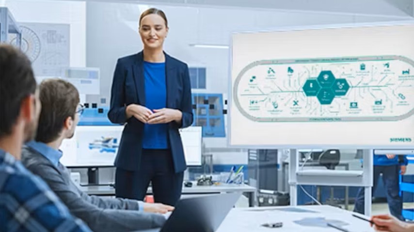 A professional business or engineering setting where a woman in a dark blazer and blue blouse is presenting to a group of colleagues.