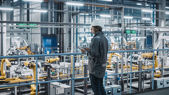 Man wearing a hardhat standing at a railing in front of machines at a manufacturing plant.