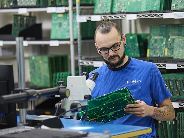 A Siemens employee inspects a circuit board, with several more circuit boards on racks behind him