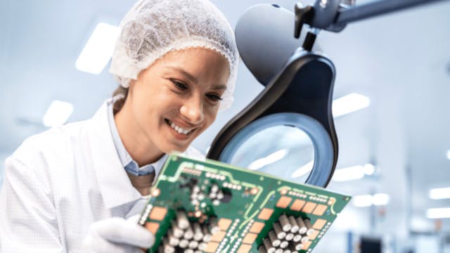 Woman in a lab looking at electronics hardware and chips under a large lab magnifying glass. 