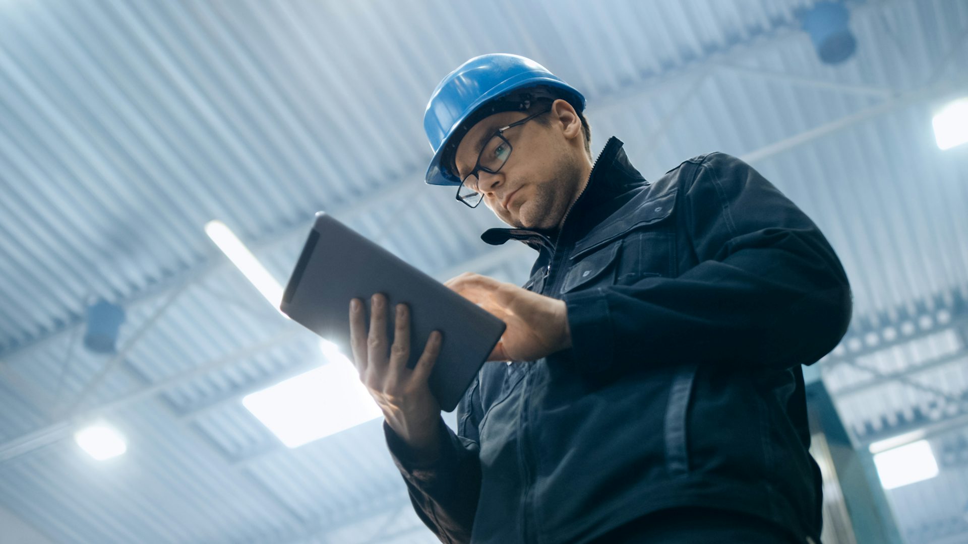A man wearing a hard hat, glasses and a jacket while looking at an iPad, checking product documentation.