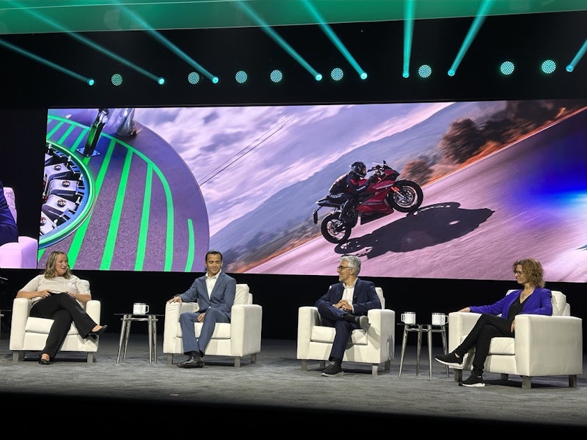 four people sit in white chairs in front of large screen at a technology conference