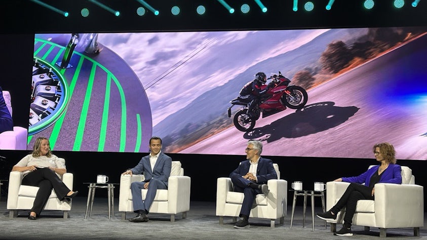 Four people sit in white chairs in front of a large screen at a Realize LIVE technology conference.
