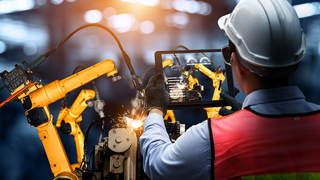A manufacturing factory worker in a white hard hat and safety vest holds a tablet recording data from robotic arms welding metal. 