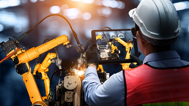 A manufacturing factory worker in a white hard hat and safety vest holds a tablet recording data from robotic arms welding metal.