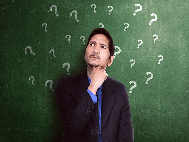 Man with thoughtful look upwards standing in front of a chalkboard, on which several question marks are written.