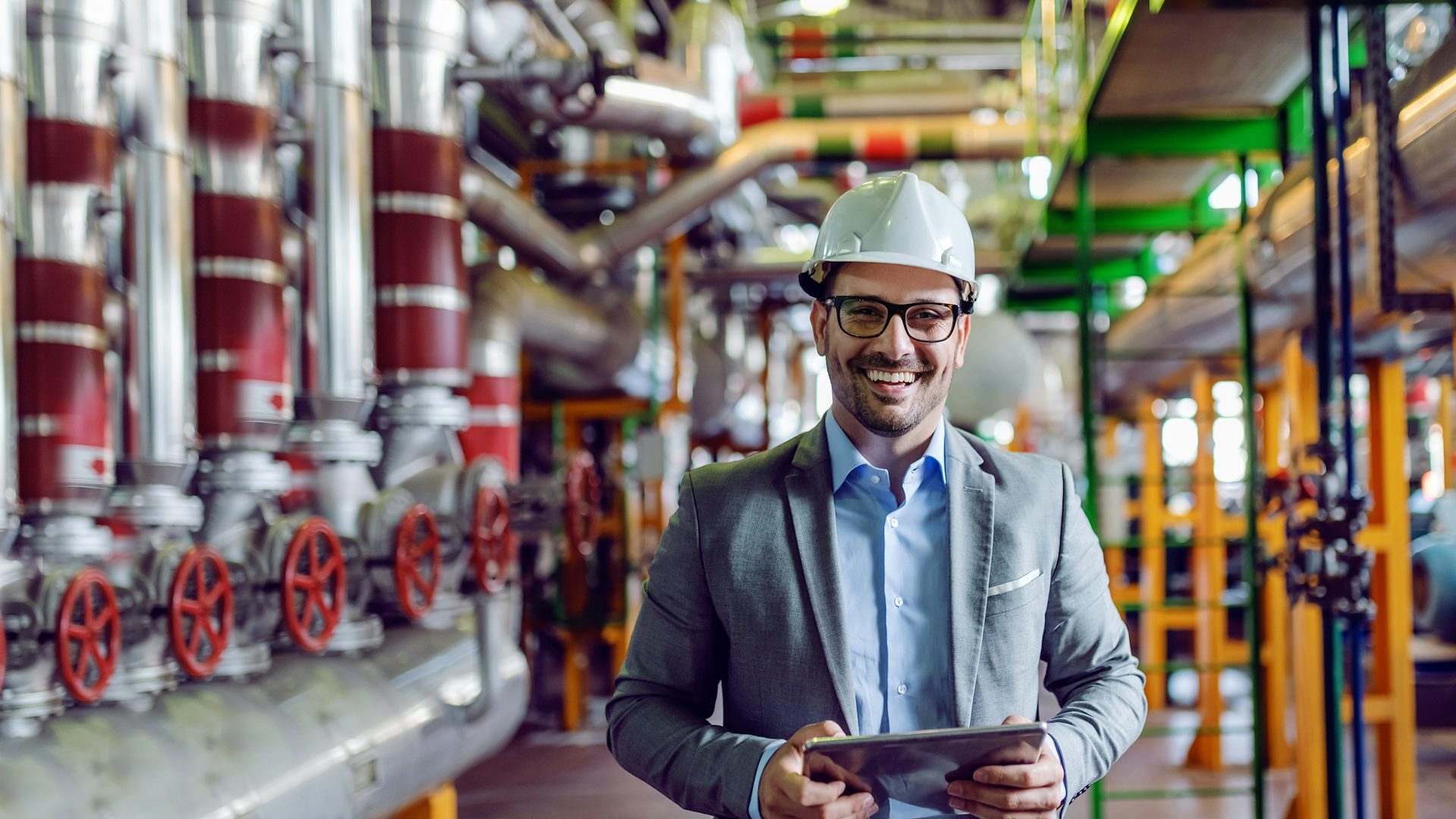 Man in a business suit wearing a hard hat smiling and standing in front of factory pipes.