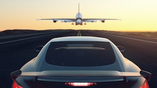 A rear view of a car driving down the middle of a two-lane road behind a plane flying low into an orange horizon. 