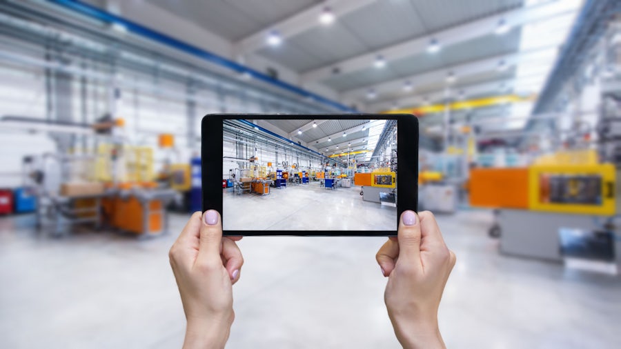 Engineer on the manufacturing floor holding a tablet in front of their face. the image on the tablet is the same manufacturing floor.