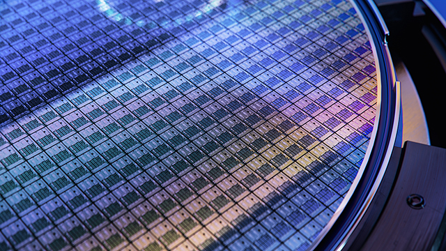 Close-up view of a silicon wafer displaying iridescent blue and purple reflections across its surface. The wafer shows a precise grid pattern of integrated circuits with metallic edge visible at bottom right.