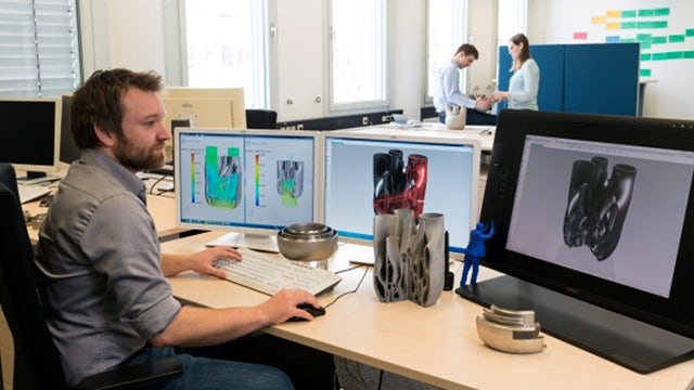 A man working with multiple screens with NX CAD open; the screens show a designed part for additive manufacturing
