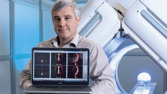 A man in front of an MRI machine holds a laptop with imaging facing the camera.