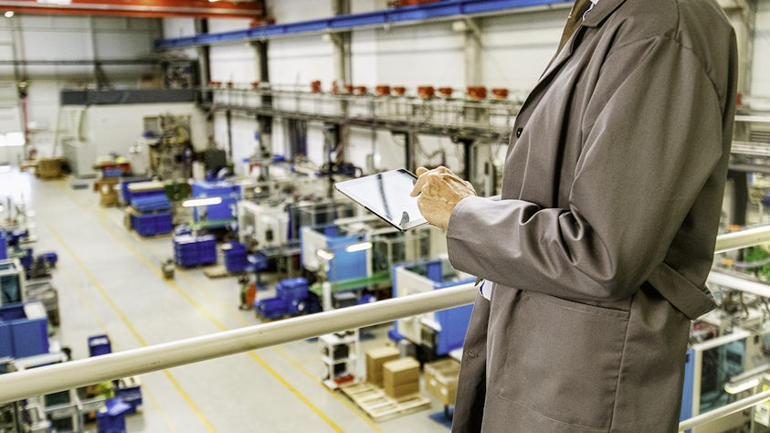 Man in a coat standing on a raised platform overlooking a factory floor, using a tablet to monitor operations on the floor.