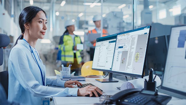 Mitarbeiter nutzt auf Qualitätsmanagement ausgelegte Konstruktionssoftware auf zwei Monitoren in einer industriellen Umgebung mit Mitarbeitern mit Sicherheitswesten im Hintergrund.