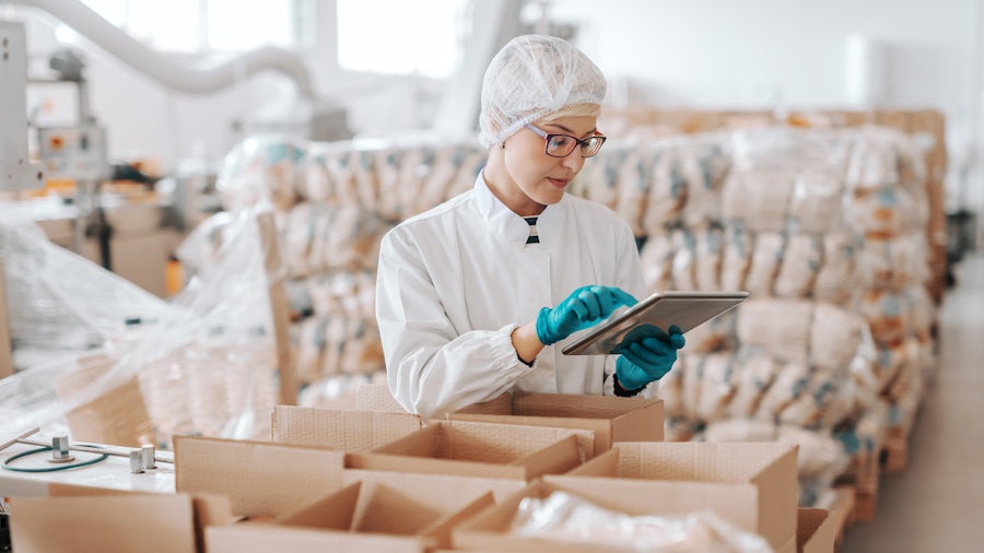 A woman in a factory wearing protective gloves and a hair net while holding and looking at a tablet.