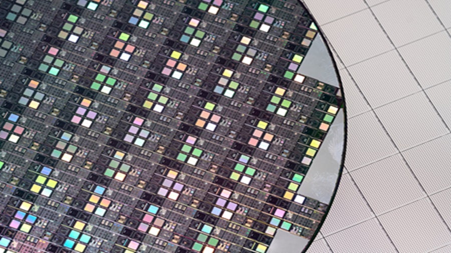 A close-up image of a silicon wafer featuring an array of integrated circuits (ICs). The wafer displays reflective, colorful patterns from the ICs, with part of the wafer extending beyond a blank grid background, highlighting its detailed design.