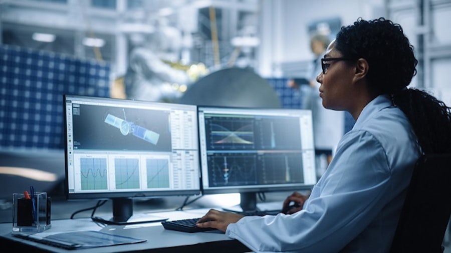 An engineer in an aerospace facility sits at desk with dual computer monitors using an MBSE solution with a satellite model and its associated data.