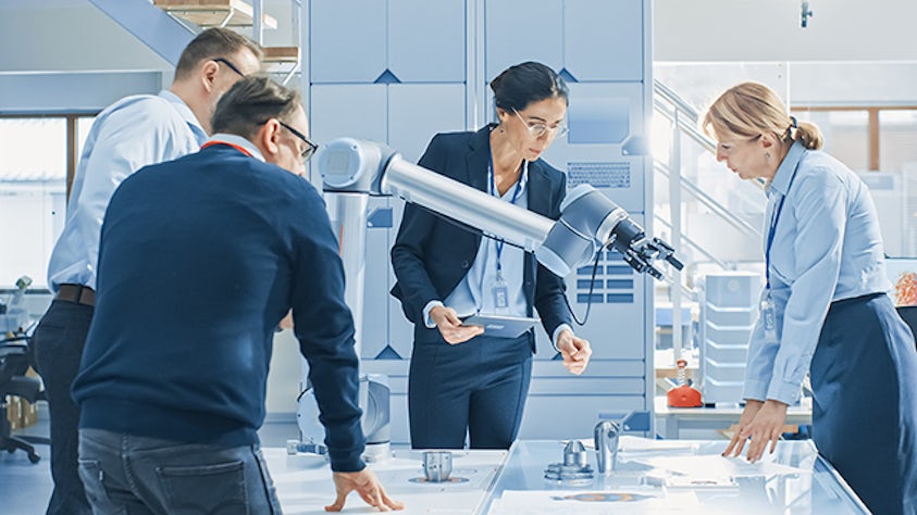 Engineers stand around a table inspecting a prototype robotic arm