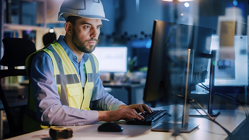 A man works on a laptop inside a manufacturing facility.