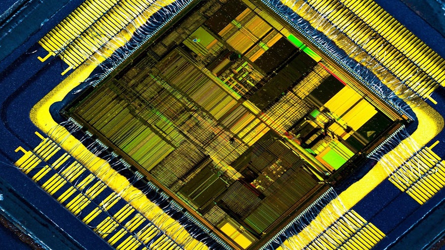 Close-up, high-magnification image of a microchip die showing detailed circuitry, gold wire bonds, and metallic connection pads around the perimeter; intricate patterns highlight the complex internal structure and layered design of an integrated circuit.