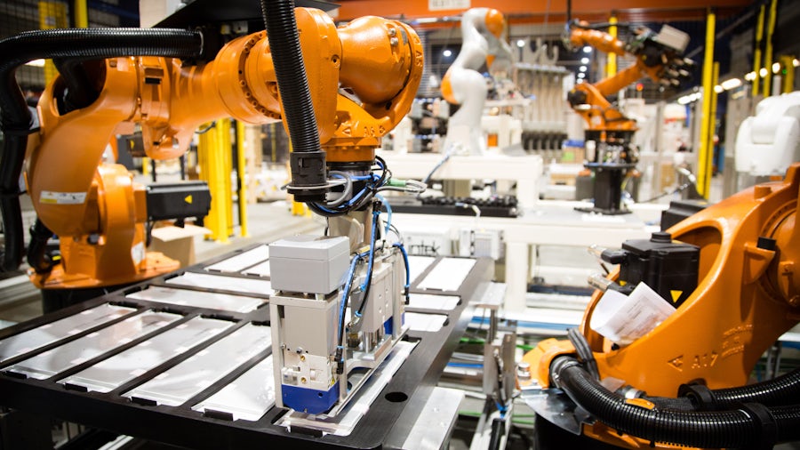 A close-up image of a shop floor with orange battery machines assembling white electric batteries.