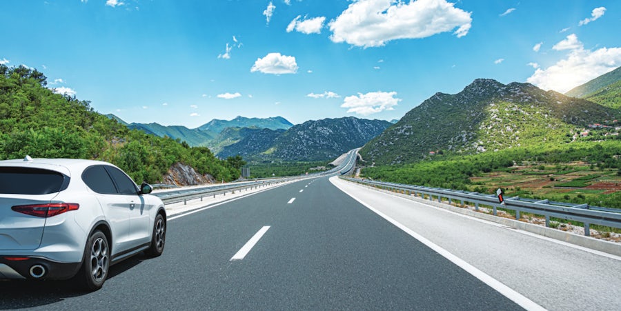  A white car travels along a highway, with mountains rising in the background under a clear blue sky.