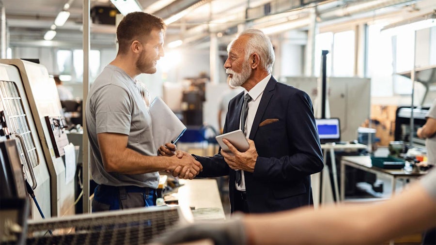 Two machine shop workers shake hands on the production floor next to CNC machines.