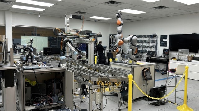 Industrial workspace featuring multiple robotic arms around a conveyor belt system, with computer monitors, workstations, and yellow safety barriers.