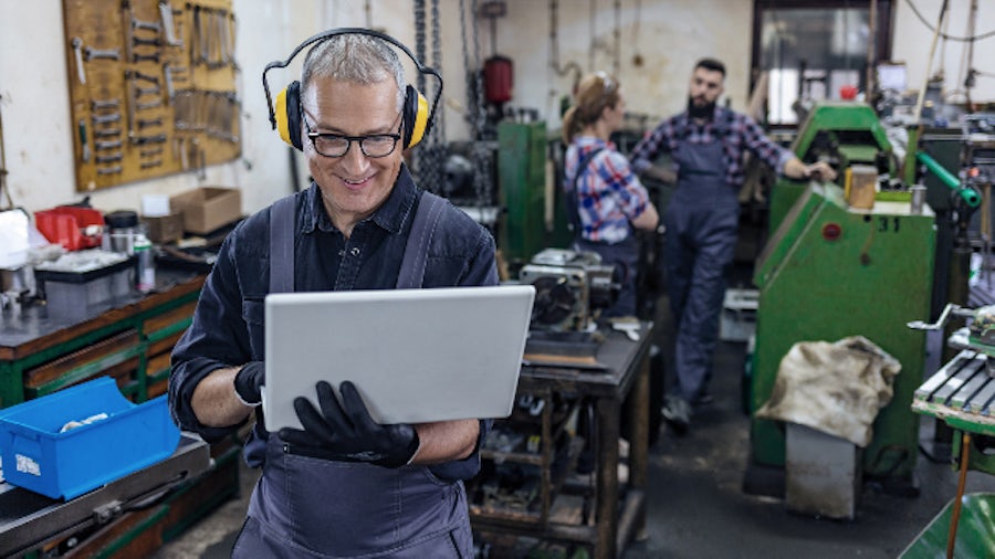 A man in a machine shop holds a laptop using technology solutions to help his small business.