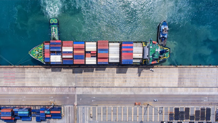 A cargo ship loaded with containers is docked at a port, with a tugboat nearby in the water.